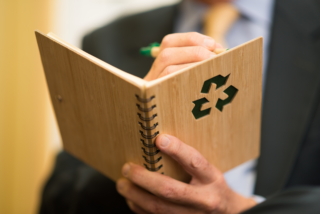 A person in a suit writing in a notebook with a green recycling symbol on the cover, emphasizing sustainability.