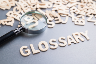 Magnifying glass highlighting wooden letters spelling “GLOSSARY” on a gray surface, surrounded by scattered wooden letters.